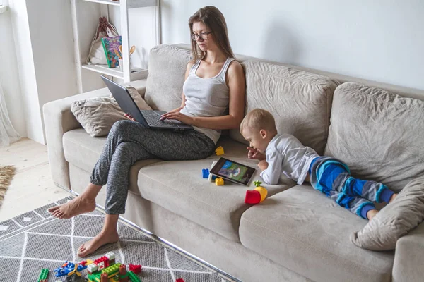 mom works at home sitting on couch with laptop while her little