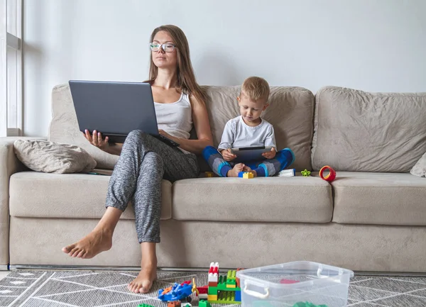 mom works at home sitting on couch with laptop while her little