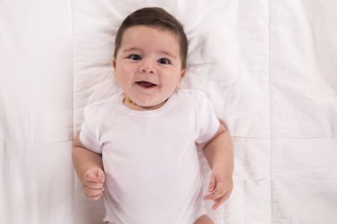 Üstten görünüm ve yakın çekim portre sevimli bebek çocuk kameraya bakarak yatak beyaz beden giyiyor. Rahatlatıcı ve çocuk odasında yatakta gülümseyen yeni doğan çocuk