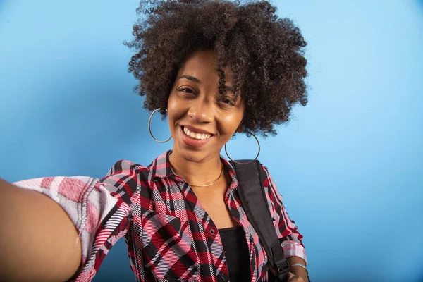 Primer plano retrato de la joven sonriente atractiva mujer brasileña africana sosteniendo teléfono inteligente, tomando foto selfie en el fondo azul . — Foto de Stock