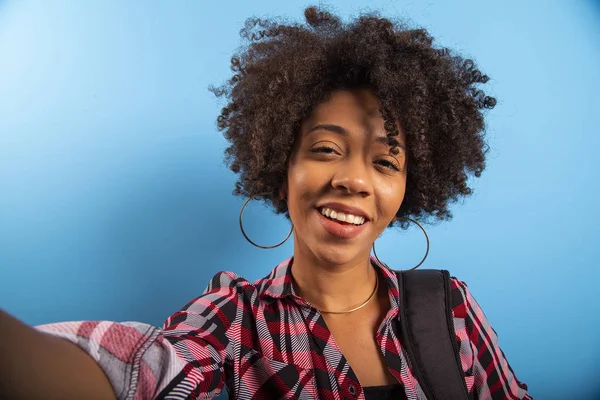 Primer plano retrato de la joven sonriente atractiva mujer brasileña africana sosteniendo teléfono inteligente, tomando foto selfie en el fondo azul . — Foto de Stock