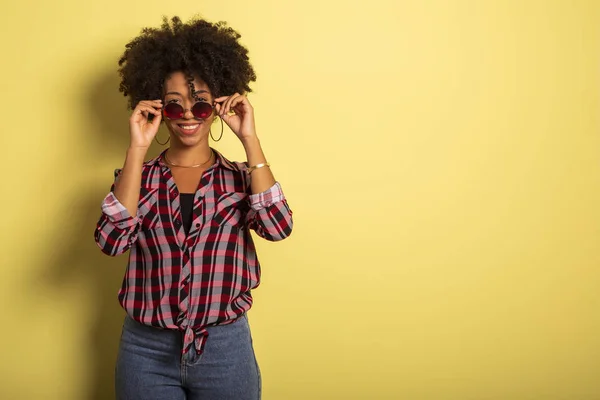 Chica brasileña afro atractiva alegre disfrutando de la vida usando gafas de sol Sobre fondo amarillo de la pared - Imagem . — Foto de Stock
