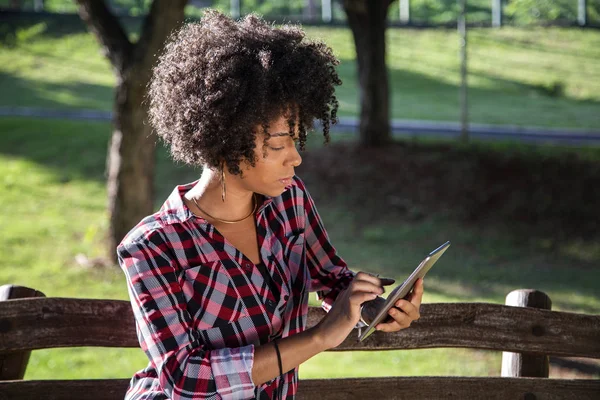 Joven hermosa brasileña con pelo negro rizado Afro con tableta, mientras está sentada al aire libre en el banco de madera en un parque — Foto de Stock