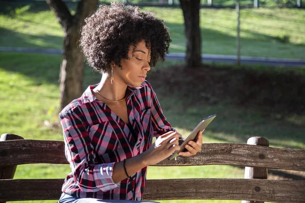 Joven hermosa brasileña con pelo negro rizado Afro con tableta, mientras está sentada al aire libre en el banco de madera en un parque — Foto de Stock