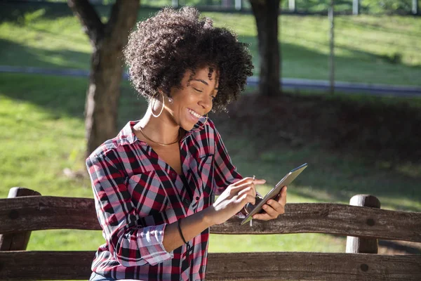 Joven hermosa brasileña con pelo negro rizado Afro con tableta, mientras está sentada al aire libre en el banco de madera en un parque — Foto de Stock