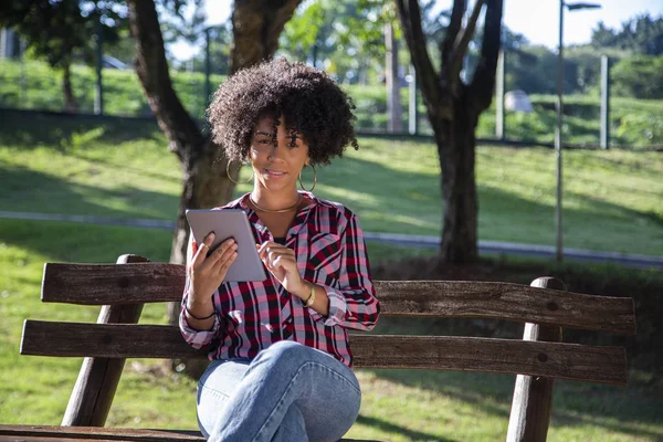 Joven hermosa brasileña con pelo negro rizado Afro con tableta, mientras está sentada al aire libre en el banco de madera en un parque — Foto de Stock