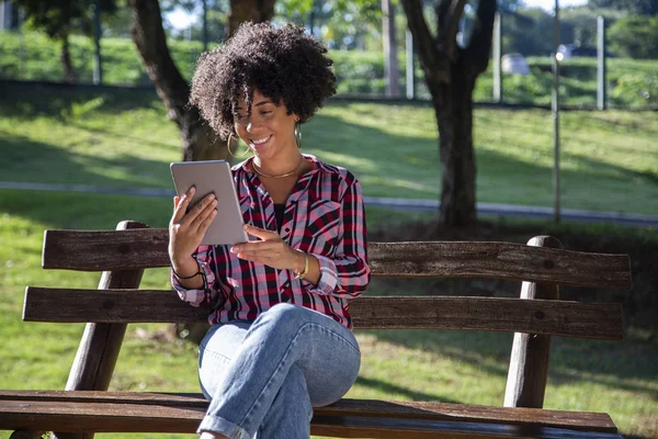 Joven hermosa brasileña con pelo negro rizado Afro con tableta, mientras está sentada al aire libre en el banco de madera en un parque — Foto de Stock