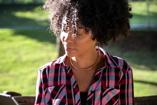 Joven hermosa brasileña con pelo negro rizado Afro con tableta, mientras está sentada al aire libre en el banco de madera en un parque — Foto de Stock