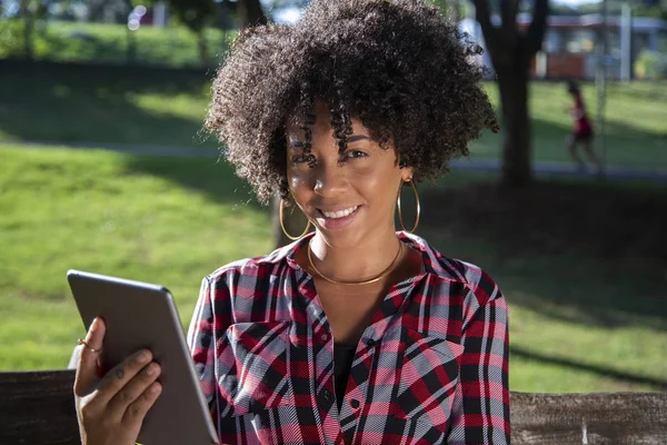 Joven hermosa brasileña con pelo negro rizado Afro con tableta, mientras está sentada al aire libre en el banco de madera en un parque — Foto de Stock