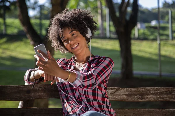 Retrato de cerca de una joven y atractiva mujer afroamericana sonriente sosteniendo un teléfono inteligente, tomando una foto de selfie y parándose en el parque con plantas borrosas en el fondo — Foto de Stock