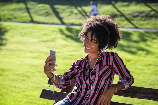 Retrato de cerca de una joven y atractiva mujer afroamericana sonriente sosteniendo un teléfono inteligente, tomando una foto de selfie y parándose en el parque con plantas borrosas en el fondo — Foto de Stock