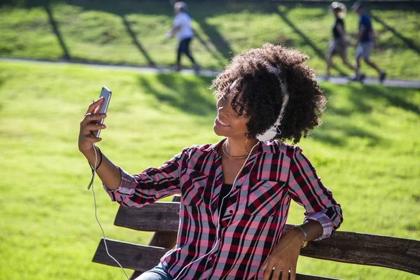 Retrato de cerca de una joven y atractiva mujer afroamericana sonriente sosteniendo un teléfono inteligente, tomando una foto de selfie y parándose en el parque con plantas borrosas en el fondo — Foto de Stock