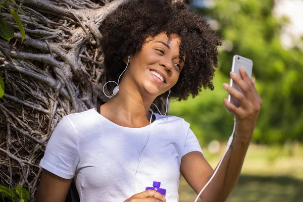 Retrato de cerca de una joven y atractiva mujer afroamericana sonriente sosteniendo un teléfono inteligente, tomando fotos de selfies y parándose en el parque con plantas borrosas en el fondo - Imagem . — Foto de Stock