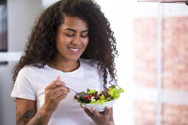 Wesoły młodych afro american kobieta jedzenie sałatka jarzynowa w domu kuchnia. — Zdjęcie stockowe