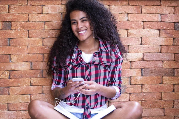Mujer estudiante feliz con peinado afro sentado con fondo de pared de ladrillo utilizando el teléfono celular y sonriendo — Foto de Stock