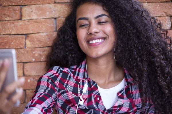 Estudiante joven afroamericana haciendo foto selfie en la cámara frontal del smartphone para compartir en redes sentadas en el suelo con fondo de ladrillo - Imagen . — Foto de Stock