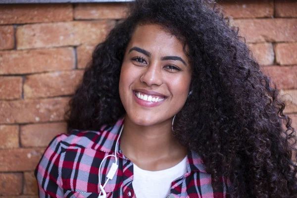 Happy student woman with afro hairstyle sitting with brick wall background using cell phone and smiling