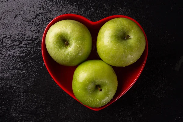 Ripe green apples into heart bowl on table close up