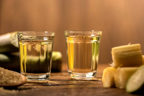 Dois copos de cachaça de ouro brasileiro com açúcar e cana-de-açúcar isolados em fundo rústico de madeira . — Fotografia de Stock