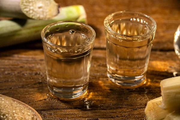 Two shot glasses of Brazilian gold  cachaca with sugar and sugarcane isolated on rustic wooden background. — Stock Photo, Image