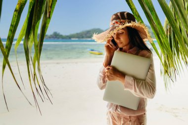 Sahilde dizüstü bilgisayar ile telefonda konuşurken bir kadın. Uzaktan seyahat ederken çalışma. Yaz tatili de Seyşeller.