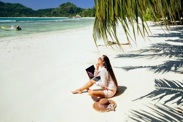 Remote working woman on beach in tropics. Working remotely on the laptop computer through the internet. Working while travelling. Summer vacation in Seychelles.