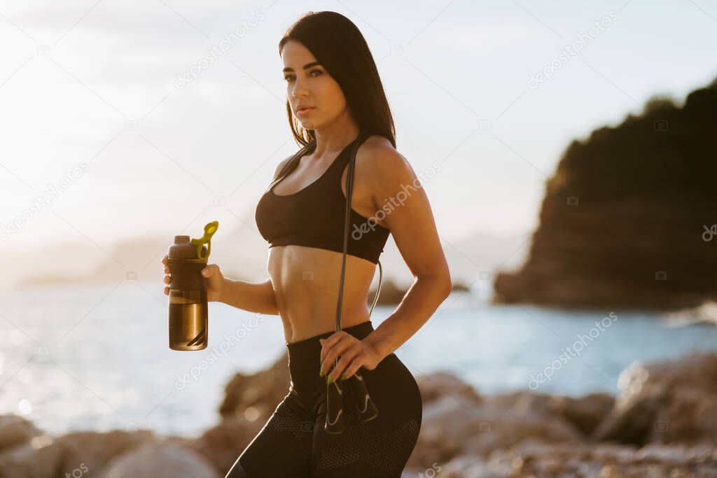 sexy athletic woman personal instructor. Beautiful sporty woman holding protein shaker and jump rope. Naked stomach abs. Fitness workout outdoors.