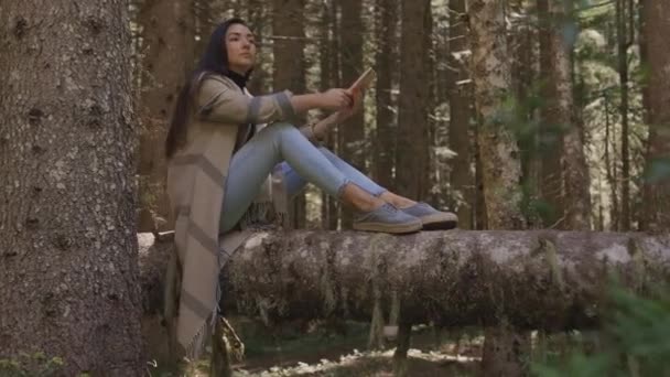 Mooie vrouw met behulp van telefoon in het bos zitten op houten log — Stockvideo