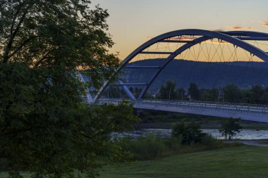 Çağdaş kemer köprüsü, ağaçlar, tepeler ve ılık günbatımı gökyüzü ile çerçevelenmiş bir nehir boyunca uzanır..