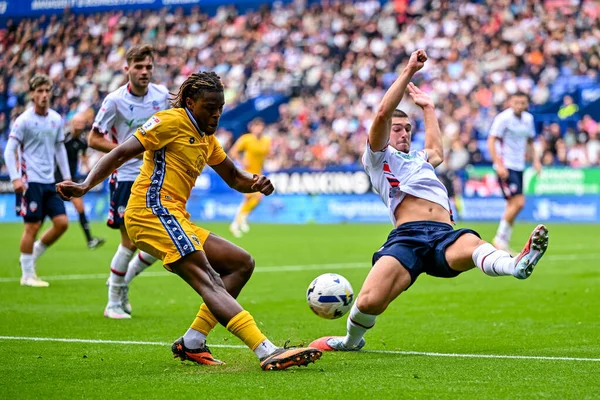 AFC Wimbledon takımından Nathan Asiimwe, Bolton Wanderers ile AFC Wimbledon arasındaki 6 Eylül 2025 'te oynanan maçta Bolton Wanderers takımından Xavier Simons' a meydan okurken bir haç taktı.
