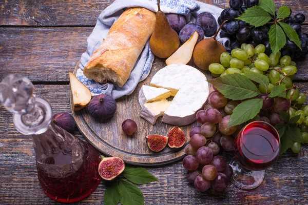 The set of products - cheese, grapes, figs, plums, pears, white bread on wooden background. Crystal decanter with red wine and glass of red wine.