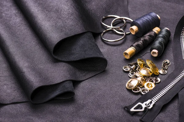 Sheets of leather for sewing leather things, a set for sewing: threads, scissors, zipper-fasteners, metal rivets, buttons on a black leather background.