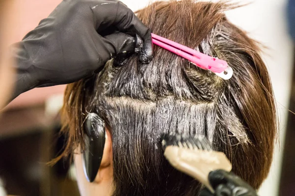 Closeup of hair dresser applying chemical color dye onto hair of customer in salon