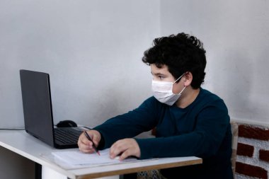 Beyaz çocuk bilgisayar ile çalışıyor ve solunum koruma maskesi takıyor.
