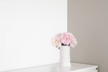 Beyaz ve gri duvar odadaki açısı. Şifonyerin üstünde beyaz vazo pembe çiçekler peonies çiçek açmış duruyor. Duvarlar iki renklerde boyanmış. Basit minimalist iç. Metin için yer.