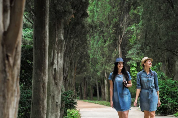 İki genç güzel esmer kız kot elbise ve şapka park uzun yeşil ağaçlar ile yol boyunca yürüyüş.