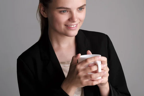 Krásná mladá dívka v černé bundě na šedém pozadí má bílý šálek kávy. — Stock fotografie