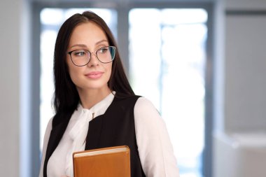 Kız öğrenci ya da öğretmen ofiste. Gözlük ve kostüm, bir defter elinde.