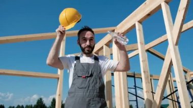 Adam Oluşturucusu Sarı kask ve gri tulum ve beyaz bir T-shirt çerçeve evin çatısı kenarına oturur. O şişe kafasına su dökülen.