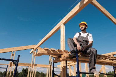 Adam Oluşturucusu Sarı kask ve gri tulum çerçeve evin çatısı kenarına oturur. Mavi gökyüzü ve açık güneşli bir gün.