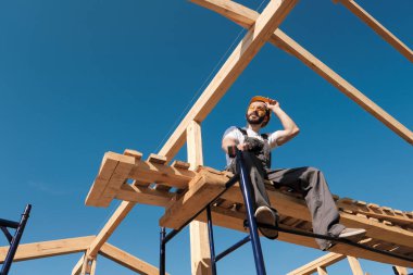 Adam Oluşturucusu Sarı kask ve gri tulum çerçeve evin çatısı kenarına oturur. Mavi gökyüzü ve açık güneşli bir gün.