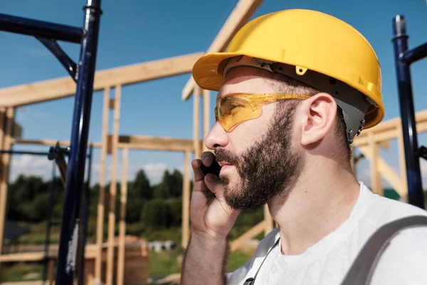 Adam Oluşturucu çatı çerçeve ev, sarı bir kask ve gri tulum arka plan üzerinde bir cep telefonu kullanır.