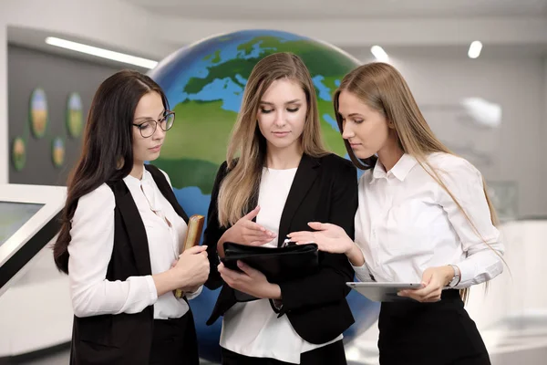 Drie mooie jonge meisjes in het kantoor met een tablet op de achtergrond van de hele wereld. Bedrijfsleven en wetenschap. — Stockfoto