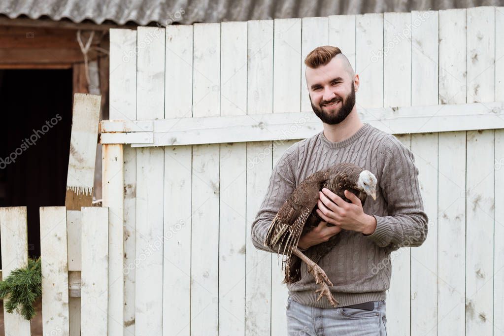 A young man with a beard, a farmer, is holding a live turkey in his hands.