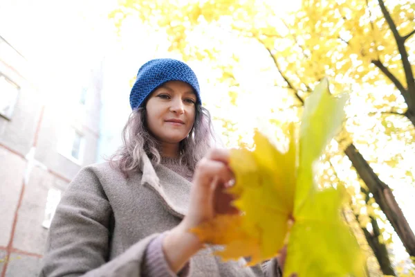 Das Mädchen in Mantel und blauem Hut auf einem Hintergrund aus Herbstbäumen und Ahornblättern. — Stockfoto