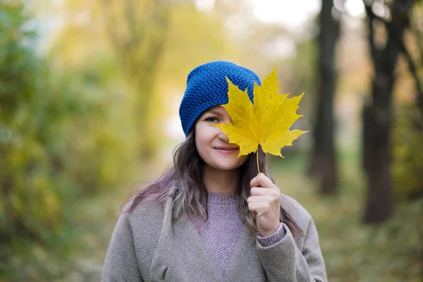 La chica en un abrigo y sombrero azul sobre un fondo de árboles de otoño y hojas de arce . — Foto de Stock