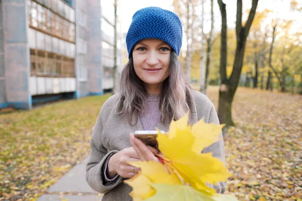 Das Mädchen in Mantel und blauem Hut auf einem Hintergrund aus Herbstbäumen und Ahornblättern. — Stockfoto