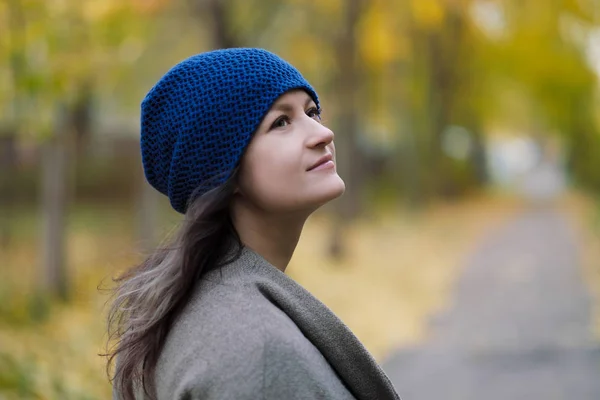 Das Mädchen in Mantel und blauem Hut auf einem Hintergrund aus Herbstbäumen und Ahornblättern. — Stockfoto