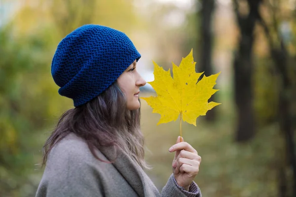 The girl in a coat and blue hat on a background of autumn trees and maple leaves. — Stock Photo, Image
