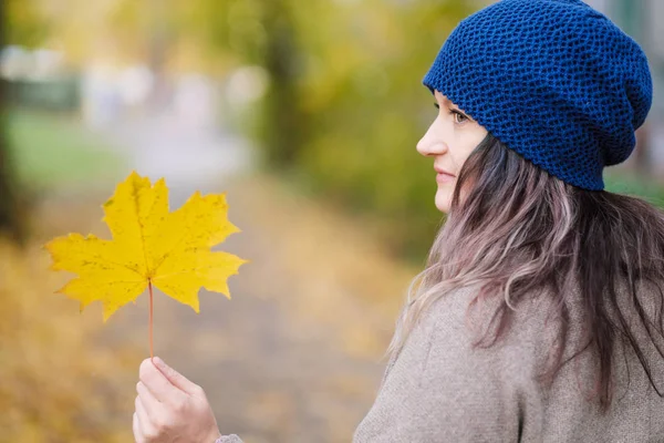 Das Mädchen in Mantel und blauem Hut auf einem Hintergrund aus Herbstbäumen und Ahornblättern. — Stockfoto